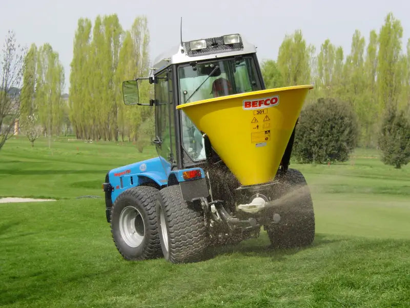 BEFCO pendulum spreader mounted on a blue tractor, distributing material on a grassy field.