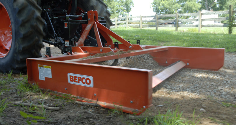 BEFCO grading scraper attached to a tractor, leveling a gravel path in a rural setting.